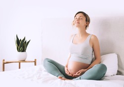 Pregnant woman in lotus pose doing meditation or breathing exercises for healthy pregnancy and preparing body for childbirth. Young expectant mother practicing yoga at home.