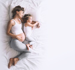 Pregnant mother and daughter resting in bed at home. Young woman with her first child during second pregnancy. Motherhood and parenting concept. Toddler girl and mom. Happy family expecting for baby.