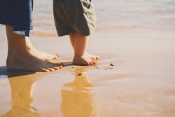 Baby's first steps. Mother and small child walking barefoot on beach sand near by ocean. Happy young family in nature. Mother and baby playing outdoors on sea beach. Close-up human feet.