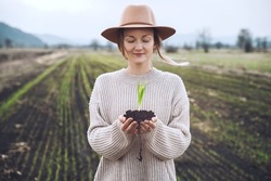 Young plant in hands in background of agricultural field area. Woman holding in hands green sprout seedling on black soil. Concept of Earth day, organic gardening, ecology, sustainable life.