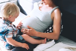 Pregnant mother and son are talking and spending time together at home. Little child boy looking at her mother pregnant tummy. Pregnancy, family, parenthood, preparation and expectation concepts.