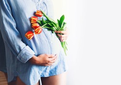 Pregnant woman with tulips flowers holds hands on belly at home interiors. Pregnancy, parenthood, preparation and expectation concept. Close-up, copy space, indoors. Tender mood photo of pregnancy