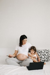 Attractive pregnant mom holding smartphone while sitting together with child daughter using laptop in white bedroom at home. Lifestyles, digital technology, togetherness, family relationships 