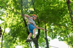 The child is a tightrope walker. Rope park. A boy teenager in a helmet walks on suspended rope ladders. Carabiners and safety straps. Safety. Summer activity. Sport. Children's playground in nature