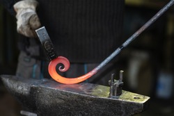 A close-up of the hands of a Blacksmith forging a curl from a red-hot flattened blank with a hammer. Handmade in the forge concept