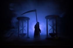 Time concept. Scary view of grim reaper silhouette standing between hourglasses with smoke and lights on a dark background. Surreal decorated picture