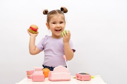 little caucasian smiling girl with toy hamburger and apple in hand is playing in store, toy cash register, vegetables, basket, scales, choice between healthy and harmful food concept