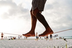 legs of a stranger performing slackline on a tight, tensioned tightrope between two palm trees in the middle of the beach overlooking the sea, sport concept