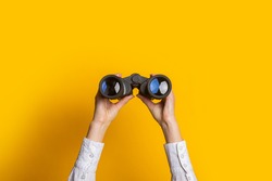 female hands hold black binoculars on a bright yellow background.