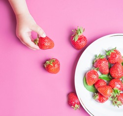 Child's hand holding strawberry on pink concrete background, plate of strawberries. Summer healthy eating concept. Top view, flat lay.