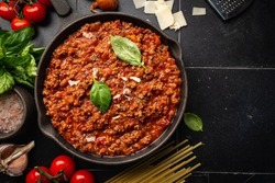 classic italian bolognese sauce stewed in a pan with ingredients on black tile background, top view