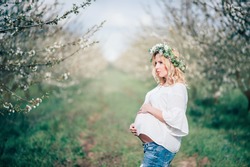 Beautiful young cheerful pregnant woman in wreath of flowers on head and jeans touching belly while walking in spring tree garden. Beauty  People Lifestyle concepts. Happy mom in waiting for baby!