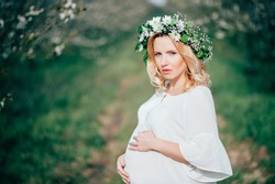 Spring coming! Beautiful young cheerful pregnant woman in wreath flowers on head touching belly while walking in spring tree garden. Beauty People Lifestyle concepts. Happy mom in waiting for baby!