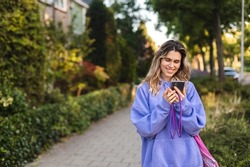 Young stylish curly blonde woman writes message on smartphone walking on green city street, hipster tourist traveling in town and planning trip using mobile phone. Look happy when read message. 