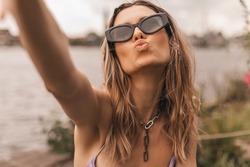 Pensive blonde woman make selfie and send blow kiss on the street near river. Outdoor shot of happy hippie lady in glasses with two thin braids and wave hair. Boho freedom style. Girl hold camera.