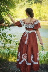 Beautiful young woman in 18-19 century dress in the forest by the pond. The romantic girl, view from the back.