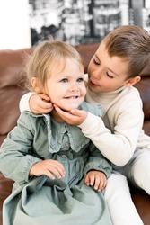 Portrait of adorable little brother and sister sitting on chair, hugging and smiling. Cute stylish dressed small kids having fun, playing together and laughing, childhood concept. High quality photo