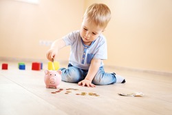 two years old child sitting on the floor and putting a euro money into a piggybank.