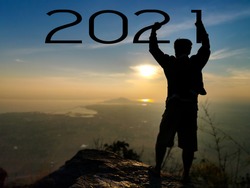 Start or end of year 2021 concept. Word 2021 floating in the air with Silhouette a man standing with  raised fist over head on the rock at the hill under the number during morning sunrise.
