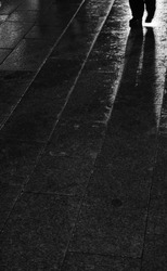 Black and white photo. City lights reflection in asphalt surface with alone (lonely) person silhouette. Rainy paving slab (road). Rain today in our town. 