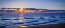 Orange sun rising in a blue cloudy sky over a flat, blue ocean. Low waves rolling into the beach. Sun rays streak across the image, lighting the sky and sea. 