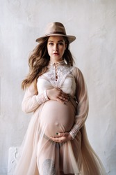Pregnancy Pretty Woman in Stylish Dress Front View Copy Space Studio Photography. Staying Girl Dressed in Style Clothing Gown and Retro Hat Caressing Belly. Beige Grunge Texture Wall on Background