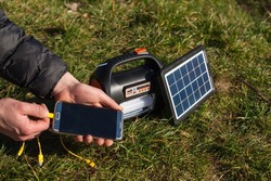 The man holds the phone in his left hand and puts it on the charger from a portable power station powered by a solar panel. A power station with a flashlight and a board powered by a solar panel.