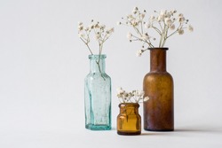 Three small glass handmade vases with white dried wild flowers. Brown and blue glass vintage bottles. Antique interior decoration objects. 