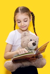 A girl is reading a book and holding a teddy bear on a yellow background. A cute girl with pigtails is learning to read and playing with her teddy bear. Children learning concept on vertical photo