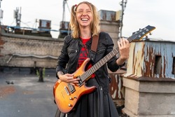 Young rock blonde girl with tattoos and two funny ponytails, laughing loudly and playing an electric guitar on the roof of an old building, front view. Musical video clip shooting. Outdoor show.