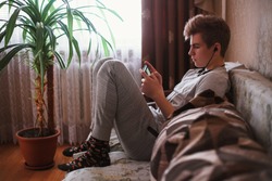 A young guy, a teenager sit on the sofa at home in headphone and writes sms on a social network or listen music on a smartphone.