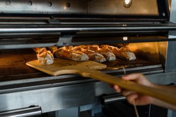 Close up of baker is taking off from oven the french baguette bread with wood peel at baking manufacture factory. French baguette bread bakery factory concept.