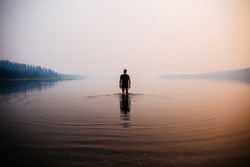Man in the water knee deep. Smokey atmosphere. Spooky. Reflection in the water. Mountains and trees in the background.