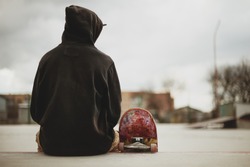 Teenager sitting in a black sweatshirt holding a skateboard on a slum background urban