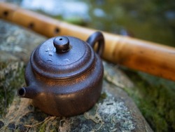 For the Chinese tea ceremony. Small brown clay teapot. Standing on a large gray stone. Against the backdrop of nature and green moss. Behind is a bamboo flute.