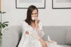 Symptoms of allergic rhinitis in women. Sick woman in white nightwear with a cold blowing her nose into a tissue paper at home. Cold weather allergies