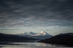 View of a mountain range covered in snow in Perth and Kinross, highlands of Scotland, United Kingdom, with snow-capped peaks reflected on the waters of the loch Tay under a dramatic sky