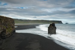A view of a volcanic beach with the waves breaking on a stone pillar near a cliff in  Reynisfjara Black Sand Beach in Vik, Iceland.