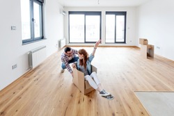 Young couple in new empty room. She is sitting on card box while he pushing her from behind