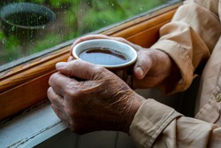 Old woman hands holding cup of tea near window with raindrops. Melancholic mood