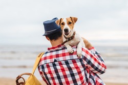 Fahionable male host of beauitiful dog, carries it on hands, come to see at sunset on seashore, spend time together, have good relationships. Unrecognizable man in checkered shirt with his pet