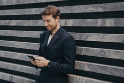Pensive young office worker in formal stylish suit standing outdoor holding mobile phone in his hands, reading last emails or messages, trying to type response on important urgent correspondence