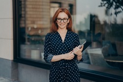 Gorgeous redhead businesswoman walks in urban place holds tablet and diary strolls near restaurant during daylight hours wears polka dot dress spectacles looks happily into distance walks in city
