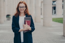 Outdoor portrait of young confident beautiful ginger woman in formal clothes holding laptop and note book and smiling at camera, waiting for business meeting, wearing spectacles, going home after work