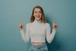 Big sale. Emotional excited amazed teen girl in casual clothes pointing up with forefingers and looking at camera with happy face expression and opened mouth, isolated over blue studio background