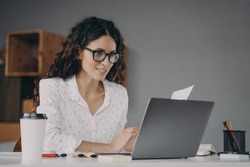 Happy european woman in glasses working on modern laptop from home, smiling attractive spanish businesswoman using computer pc while sitting at desk in office, typing on keyboard
