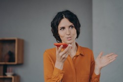Beautiful brunette Italian businesswoman elegantly speaks on loudspeaker on mobile holds phone in hand close to her face, young woman recording voice message while working remotely at home