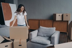 Smiling european woman is packing things. Girl moves to new house. Young lady is unpacking cardboard boxes in new apartment. Big new living room with armchair. Real estate purchase concept.