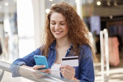 Positive female model dials number of credit card on smart phone, checks her bank account, makes shopping, needs some money for paying purchase. People, payment, technology and buying concept