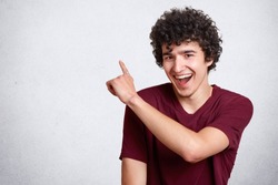 Portrait of stylish handsome young guy dresses casual outfit, with dark curly hair isolated on white background. Laughing teenager being in good mood points up. People, youth and lifestyle concept.